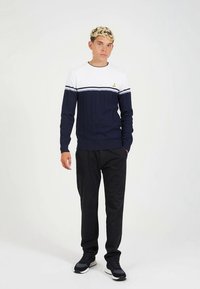 Sweater listrado em azul-marinho e branco com padrão texturizado, apresentando um emblema amarelo no peito. Combinado com calças escuras e ténis.