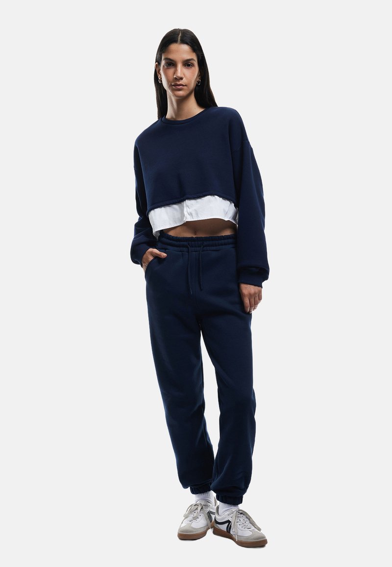 Marineblå cropped sweatshirt med et hvidt lag under, parret med løstsiddende marineblå joggers. Begge dele har en blød tekstur og et afslappet design.