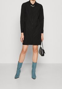 Abito camicia lungo nero con maniche lunghe, chiusura a bottoni e colletto decorativo. Abbinato a stivali alla caviglia azzurri chiari ad altezza ginocchio e una borsa nera.