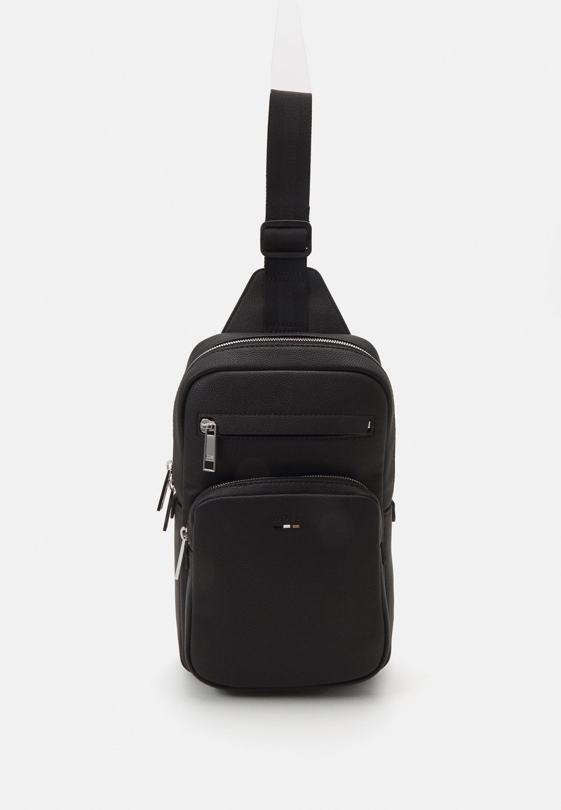 Sac à bandoulière noir en matériau texturé, avec plusieurs compartiments zippés et une sangle plate. Présente un marquage minimal à l'avant.