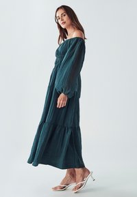 Tirkizna maksi haljina s otpalim rukavima, dugim balonskim rukavima, skupljenim strukom i slojevitim dizajnom. Uparena s bijelim sandalama na visoku petu.