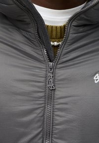 Veste isolante grise à finition texturée, dotée d'une fermeture éclair frontale et d'un détail logo ; portée sur un haut superposé vert et blanc.