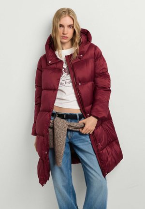 Frau trägt eine lange bordeauxrote Steppjacke mit Kapuze, weißes Grafik-T-Shirt, blaue Jeans und einen braunen Pullover um die Taille gebunden.