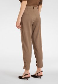 Frau trägt hoch taillierte, zulaufende braune Hose mit Knöchelriemchen, kombiniert mit dunkelbraunen Slingback-Schuhen mit niedrigem Katzenabsatz, steht auf weißem Boden.