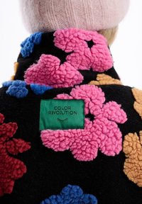 Čierna fleecová bunda s živými kvetinovými aplikáciami v ružovej, modrej a oranžovej farbe, so zelenou nášivkou "COLOR REVOLUTION" na chrbte.