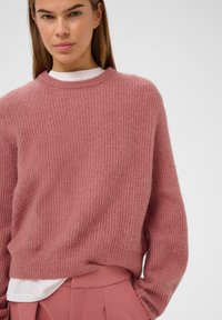Vævet lyserød sweater med et struktureret, ribbet mønster, rund halsudskæring og lange ærmer, parret med højtaljede lyserøde bukser.