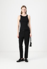 Top sans manches noir au design ajusté, associé à un pantalon noir tailleur et des chaussures lacées noires, tenant un petit sac à main noir.