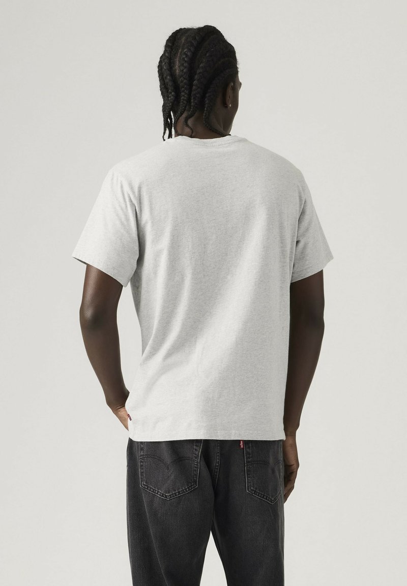 Mann mit geflochtenem Haar, der ein schlichtes, hellgraues T-Shirt mit kurzen Ärmeln und schwarze Jeans trägt, steht mit dem Rücken zur Kamera vor neutralem Hintergrund.