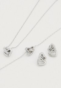 Σετ κοσμημάτων σε ασημί απόχρωση με σχέδια από διαπλεκόμενες καρδιές, διακοσμημένο με καθαρούς κρυστάλλους ριντόν. Περιλαμβάνει κολιέ και ταιριαστά σκουλαρίκια.