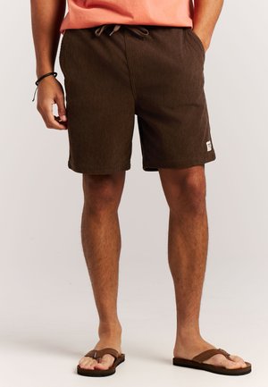 Man die bruine corduroy shorts met een trekkoord draagt en bruine slippers, staand tegen een effen achtergrond.