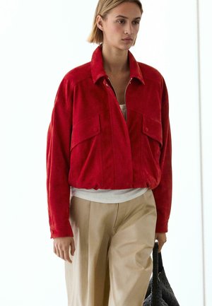 Kvinde går indendørs iført en rød lynlåstrøje, beige bukser, hvid top og holder en sort flettet taske i højre hånd.