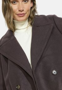 Cappotto marrone scuro con design doppiopetto, ampi risvolti e due grandi bottoni. Indossato sopra un dolcevita bianco con texture a coste.