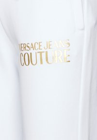Bílé tepláky s hladkým povrchem, které mají na stehně zlatě potištěné logo "VERSACE JEANS COUTURE".