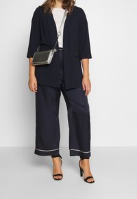 Blazer bleu marine à manches trois-quarts, associé à un pantalon large en satin bleu marine avec un liseré rose et des sandales à talons hauts noires.