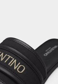 Sandale noire à glissière avec une large bride portant "VALENTINO" brodé en fil beige sur le dessus.