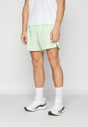 Jasnozielone spodenki sportowe z bocznymi rozcięciami, w połączeniu z białymi butami z zielonymi akcentami. Model nosi białą bluzkę i białe skarpetki.