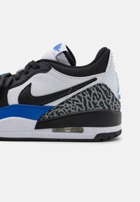 Nike sportcipők, amelyek fekete felsőrésszel, fehér és kék díszítésekkel, texturált szürke sarokkal és látható légpárnás betéttel rendelkeznek a talpban.