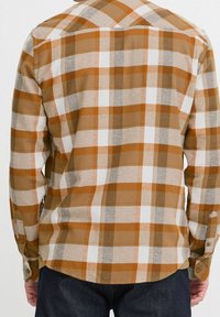 Camisa de cuadros marrón y crema, de manga larga, con textura de tela visible, botones en los puños y un dobladillo curvado. No se observan elementos gráficos.