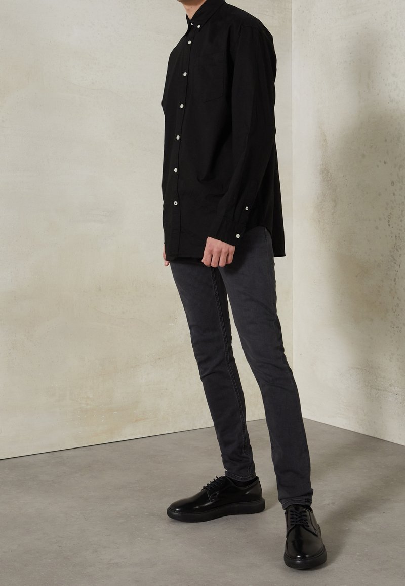 Camisa negra abotonada con mangas largas y un bolsillo, combinada con jeans ajustados gris oscuro y zapatos negros con cordones, sobre un fondo neutro.