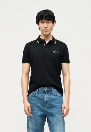 Fiatal férfi, aki fekete, sárga szegéllyel és logóval díszített pólót visel, kék farmerral, világos, egyszerű háttér előtt áll.