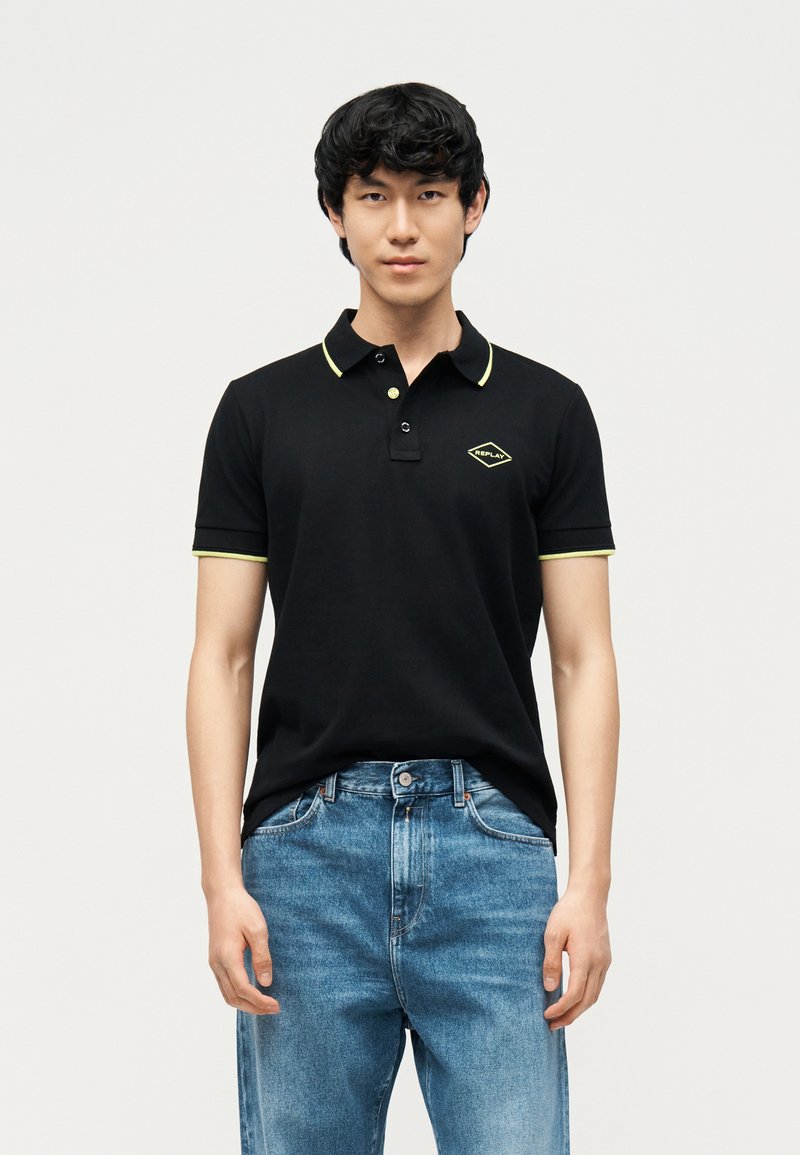 Νέος άνδρας που φοράει μαύρο πόλο μπλουζάκι με κίτρινο περίγραμμα και λογότυπο, συνδυασμένο με μπλε τζιν, στέκεται μπροστά σε ένα απλό φωτεινό φόντο.