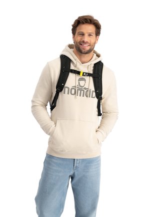 Beige hoodie met een grafisch "nomad" logo, kangoeroezak en trekkoord capuchon. Draagt over blauwe denim jeans en wordt aangevuld met een zwarte rugzak.