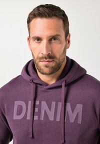 Felpa viola con grafica frontale che recita "DENIM". Presenta un cappuccio con coulisse e una texture morbida, progettata per un abbigliamento casual.