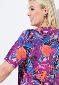 Kortärmad topp i lila tyg med ett livligt tropiskt lövtryck i orange, blått och rosa, med en avslappnad passform och rund halsringning.