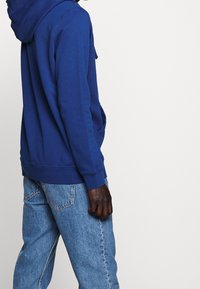 Felpa blu in cotone con tasca frontale e cappuccio regolabile con cordino, abbinata a jeans azzurri, vista di lato.
