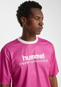 Rosa sport t-shirt i lätt material. Har en vit rund halsringning och fet vit text som säger "hummel PERFORMANCE" på framsidan.