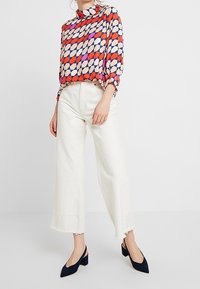 Blouse colorée avec des motifs géométriques en rouge, bleu et beige. Pantalon blanc à jambes larges et talons bleu foncé. Ourlet effiloché sur le pantalon.