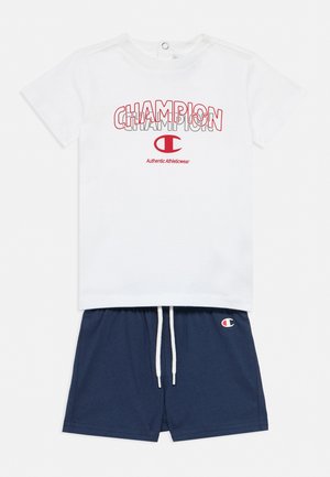 Camiseta Champion blanca de manga corta con logo y texto en rojo, combinada con pantalones cortos azul marino con cordón que presentan el emblema de Champion.