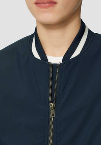 Marineblau Bomberjacke mit einem weißgestreiften Kragen, ausgestattet mit einem Metallreißverschluss und einer glatten Textur. Nahaufnahme, die die Designdetails hervorhebt.