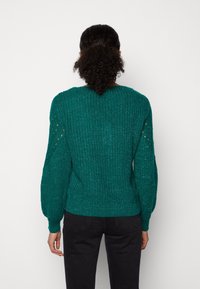 Pull en tricot bleu sarcelle avec tissu texturé, manches ballon et détails ajourés sur les épaules. Ourlet et poignets côtelés.