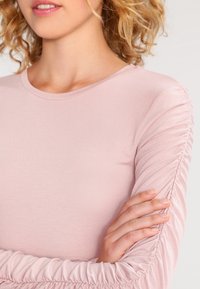 Langærmet top i blød pink stof med rund halsudskæring og samling detaljer langs ærmerne, med en glat og strækbart materiale.