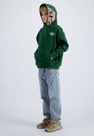 Jongen draagt een groene hoodie met een tijgergezicht op de capuchon en borst, lichte gescheurde spijkerbroek en beige sneakers, staand met handen in de zakken.