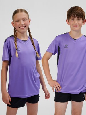 Zwei lächelnde Kinder, die identische lila Sportshirts und schwarze Shorts tragen, stehen vor einem schlichten weißen Hintergrund.