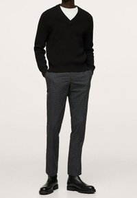 Zwarte geribbelde gebreide V-hals trui, gecombineerd met grijs geruite tapered broeken en zwarte enkellaarsjes. De outfit heeft een gelaagd uiterlijk met een witte shirt.