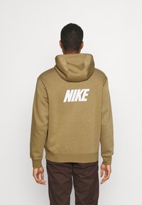 Sudadera con capucha color caqui con un gran logo blanco "NIKE" en la espalda, que presenta una textura suave, bolsillo delantero tipo canguro y capucha con cordón ajustable.