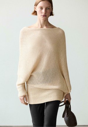Kvinde iført løs beige strikket sweater og sorte bukser, der holder en mørk vævet håndtaske mod en ensfarvet lys baggrund.