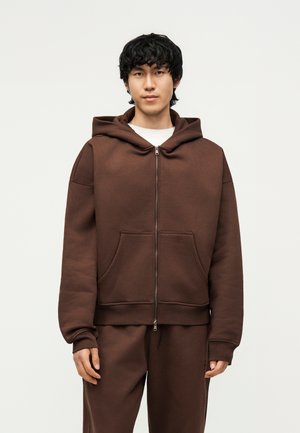 Noor mees mustade juustega, kellel on seljas pruun lukuga kapuutsiga pusa ja sobivad püksid, seisab lihtsa heleda tausta ees.