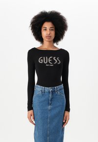 Body a maniche lunghe nera con il logo "GUESS" in strass, abbinato a una gonna di jeans a vita alta con uno spacco frontale e chiusura a bottoni.