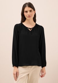 Blusa nera a maniche lunghe con scollatura rotonda, dotata di dettagli arricciati e un laccio sul davanti. Tessuto morbido con una vestibilità comoda.