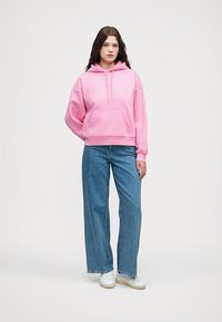 Roza dukserica s kapuljačom i prednjim džepom, vezicom i rebrastim rubom, u kombinaciji s svjetloplavim širokim trapericama i bijelim tenisicama.