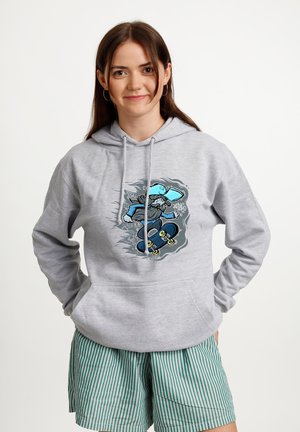 Mladá žena s hnědými vlasy, která má na sobě šedou mikinu s kapucí s obrázkem kresleného skejťáka a zelené pruhované šortky, stojí s rukama v kapse.