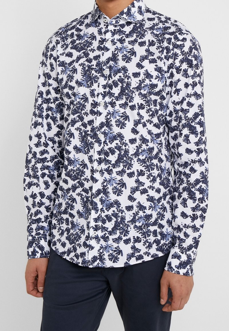 Homme portant une chemise blanche à manches longues avec un motif floral bleu foncé et un pantalon bleu foncé.