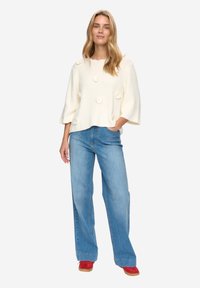 Krémový svetr s texturovanými květinovými dekoracemi, širokými rukávy; kombinován s světle modrými kalhotami širokých nohavic a červenou obuví. Jednoduchý, neformální outfit.