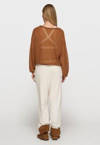 Pull en maille brun surdimensionné avec un dos transparent ; associé à un pantalon ample couleur crème et des bottes marron doublées de fourrure.