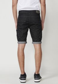 Dunkelblaue Denim-Shorts mit umgeschlagenem Saum, Gesäßtaschen und einem Etikett am Bund. Getragen mit schwarzen Sneakers. Glatte Textur, taillierte Passform.