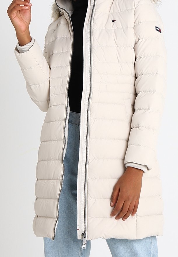 Lätt, beige pufferjacka med horisontella sömmar, hög krage, dragkedjestängning och pälsfodrad huva. Har subtila logotypdetaljer.